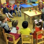 Children sit in a semicircle, teacher and girl in the center